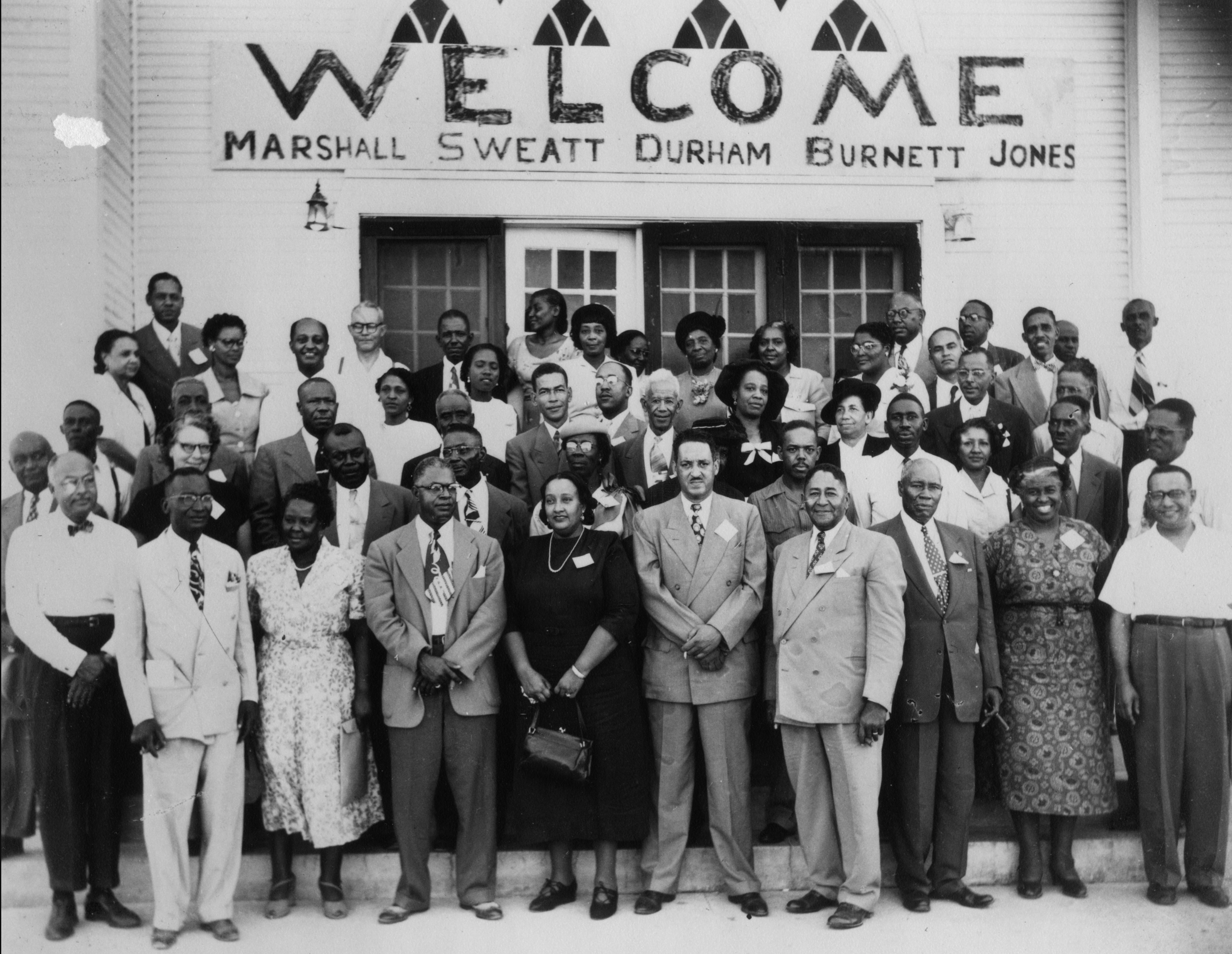 Forty African Americans in business attire pose on the steps of building festooned with a banner that reads "Welcome Marshall, Sweatt Durham, Burnett, Jones